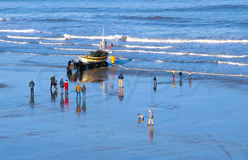 Comp image : ea00416 : A few spectators watch a fisherman bring his boat ashore at Cromer, on the North Norfolk coast of England, on a sunny winter day. The sea and wet foreshore reflect the blue sky.