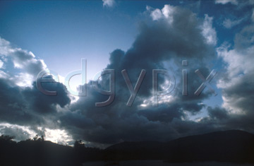 Comp image : sky0113 : Dramatic billowing dark clouds covering the bright sun in a blue sky, over a heavy land mass on the skyline