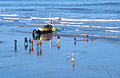 A few spectators watch a fisherman bring his boat ashore at Cromer, on the North Norfolk coast of England, on a sunny winter day. The sea and wet foreshore reflect the blue sky.