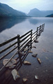 Morning autumn sun and reflection of a fence and rocks in the still surface of Buttermere lake, in the English Lake District, with Fleetwith Pike in the distance