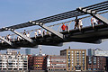 People walking in sunshine on the Millennium Bridge over the River Thames in London, England, with buildings on the north bank of the river in the backgound