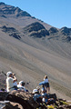 A resting group of trekkers is dwarfed by the bulk of Jebel Rhat, a mountain in the High Atlas mountains of Morocco; a massive field of scree in the mid distance