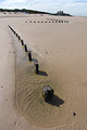 A zigzag line of wooden posts in sunshine on a deserted sandy beach at low tide on the flat North Norfolk coast of England, under a clear blue sky