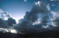 Dramatic billowing dark clouds covering the bright sun in a blue sky, over a heavy land mass on the skyline