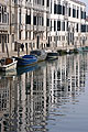 Long reflections of grey buildings lining a canal in the Cannaregio area of Venice, Italy, with a line of boats in the mid distance